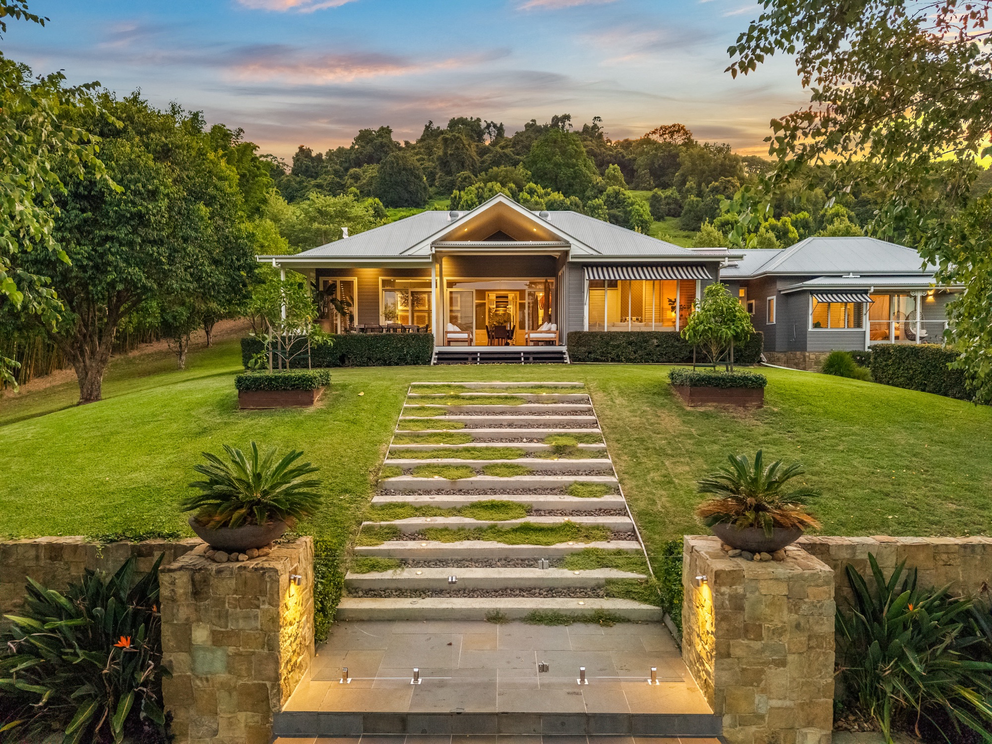 Grand front approach at twilight with stone steps, garden lighting and illuminated homestead